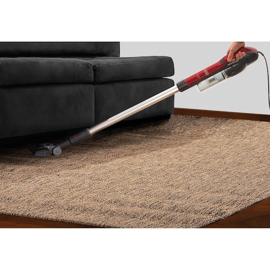 BLACKDECKER AVT12 stick vacuum with long handle cleaning under black sofa on brown rug.