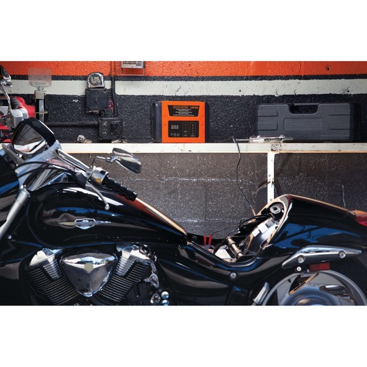 BLACKDECKER BC12 battery charger on wall shelf beside toolbox, motorcycle parked below in garage setting.