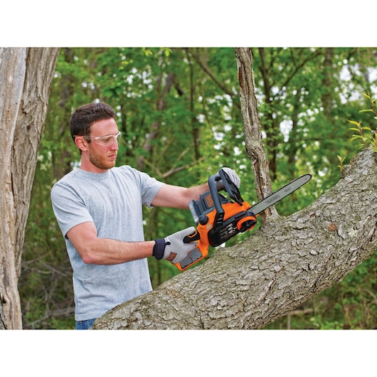 Man using BLACKDECKER BCCS1020D1 cordless chainsaw to cut tree branch outdoors, wearing work gloves.