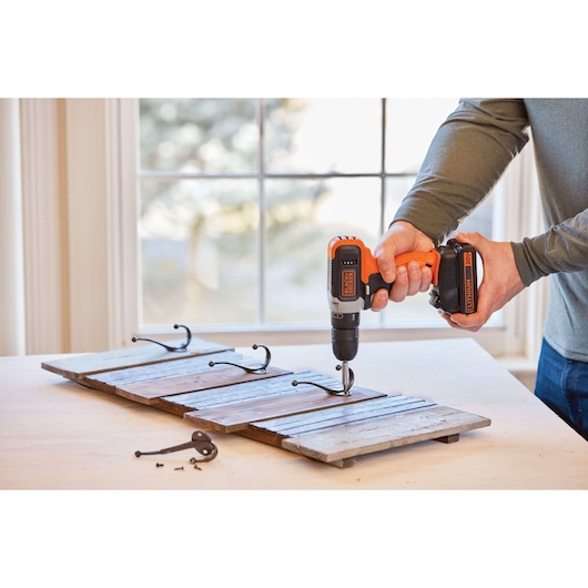 BLACKDECKER BCD70250PK cordless drill used to attach metal hooks to wooden planks, indoor table setting.