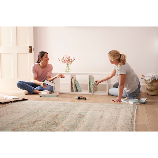 BLACKDECKER BCRTA601I cordless screwdriver on floor with two women organizing books on a white shelf in living room.