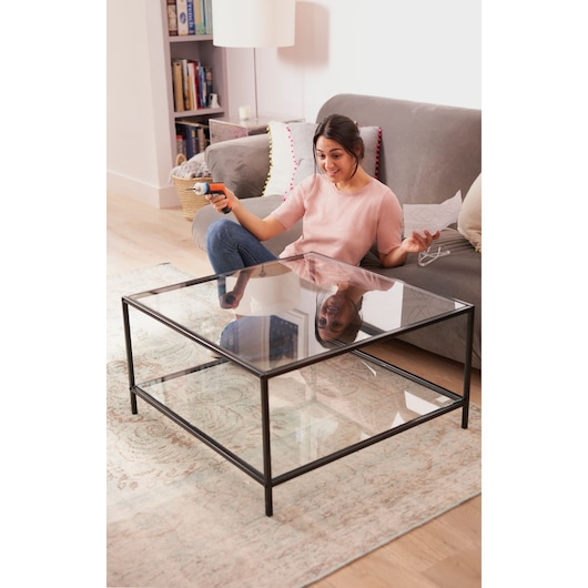 Photo of a person using a BLACKDECKER BCRTA601I cordless screwdriver while assembling a glass coffee table in a living room.