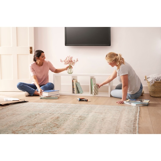 BLACKDECKER BCRTA601I cordless screwdriver on floor in living room near two people assembling white bookshelf.