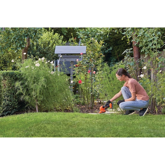 Person using BLACK+DECKER BCSS820D1 cordless shrub and grass shear in a garden near flowers and bushes.