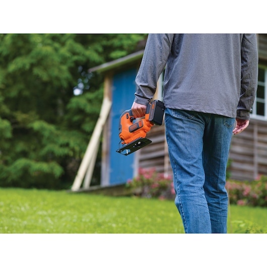Photo of BLACKDECKER orange cordless jigsaw BCK22S1S held by person outdoors near a wooden shed.