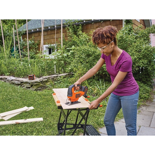 BLACKDECKER BCK22S1S jigsaw being used outdoors on plywood on a portable workbench, with grass and plants nearby.