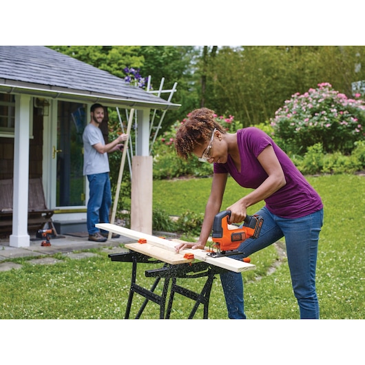 Photo of BLACKDECKER BCK22S1S jigsaw in use outdoors, cutting wood on a workbench, gardening shed in background.