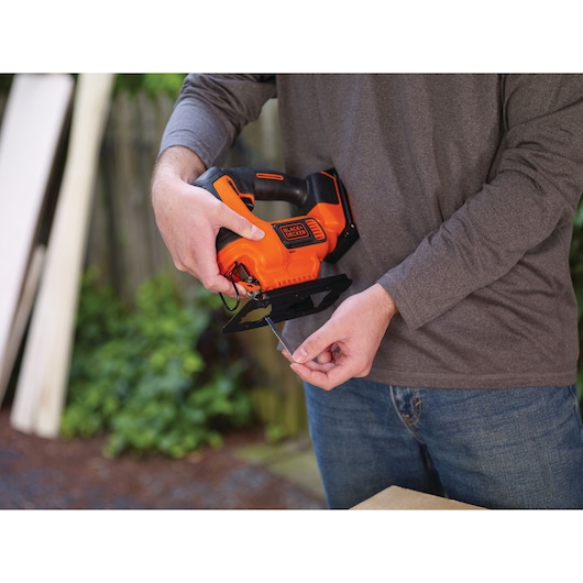 Person holding BLACK+DECKER BCK22S1S cordless jigsaw, inserting blade, outdoors with wood in background.