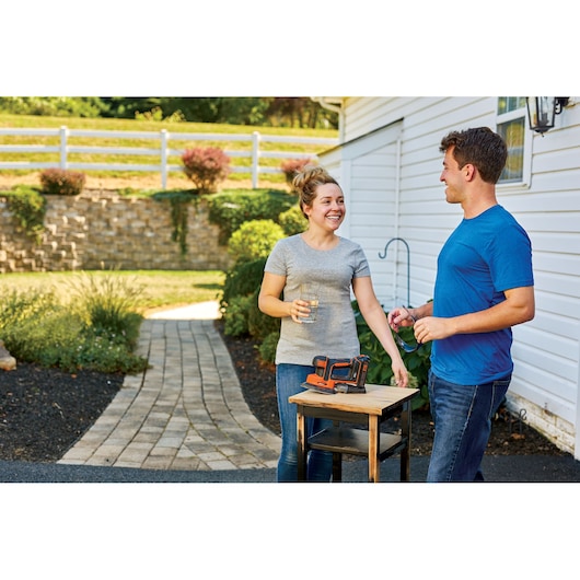 BLACKDECKER BCK22S1S power tool on small wooden table outdoors, two people standing nearby, garden and house in background.