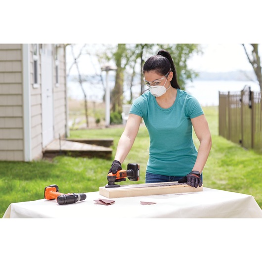 A person using a BLACK+DECKER cordless power tool on a piece of wood outdoors, with another power tool and sanding sheets visible on the table. The person's face is blurred for privacy.