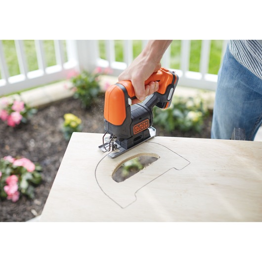 A person uses a BLACK+DECKER cordless jigsaw to cut a shaped outline in a piece of plywood, outdoors near a garden.