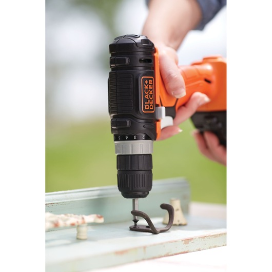 A person uses a BLACK+DECKER cordless drill to attach a metal hook to a wooden surface.
