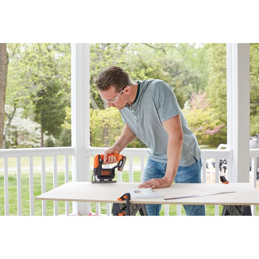 A person using a BLACK+DECKER cordless jigsaw to cut a piece of wood on a table outdoors. The wood is secured with clamps, and the scene shows a porch with greenery in the background. The person's face is blurred.