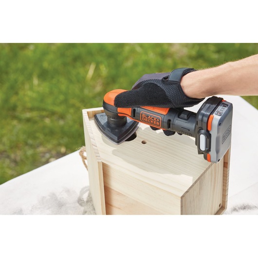 A person wearing gloves is using a BLACK+DECKER cordless power tool to sand the surface of a wooden box outdoors.