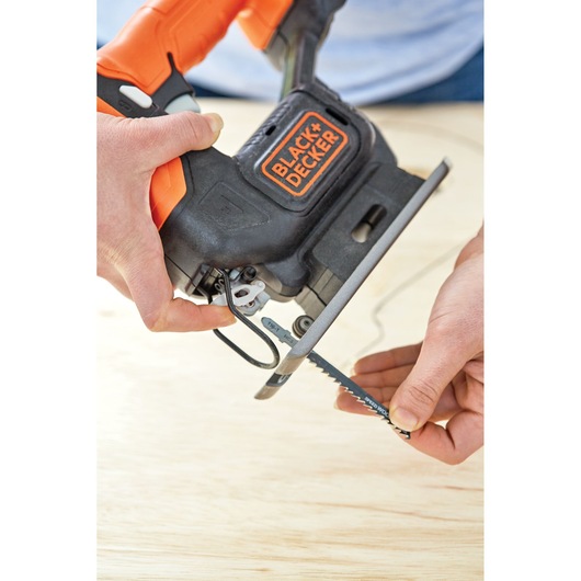 A close-up of hands installing a saw blade into a BLACK+DECKER jigsaw, with an orange and black handle, over a light wooden surface.