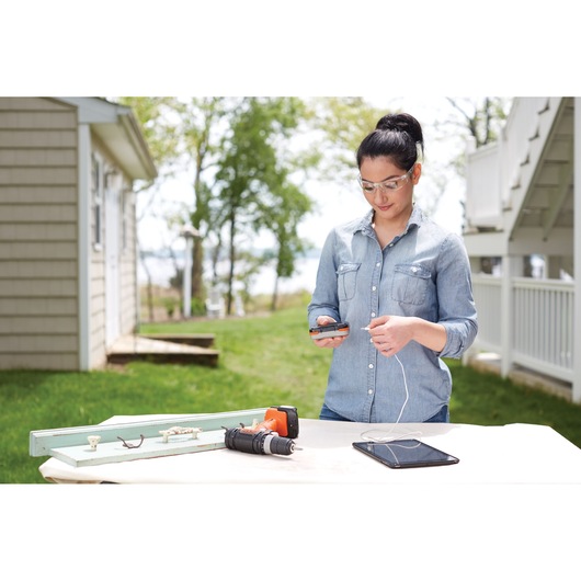 A person standing outdoors next to a table with a BLACK+DECKER cordless drill, electronic devices, and woodworking materials. The person is holding a mobile device with a cable attached to it, possibly charging or transferring data. The setting includes a house and greenery in the background.