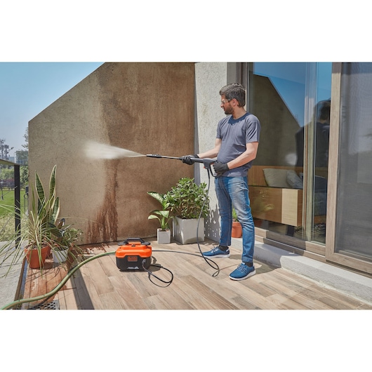 BLACKDECKER BEPW1600H pressure washer cleaning outdoor wall, orange unit on patio with hose and plants.