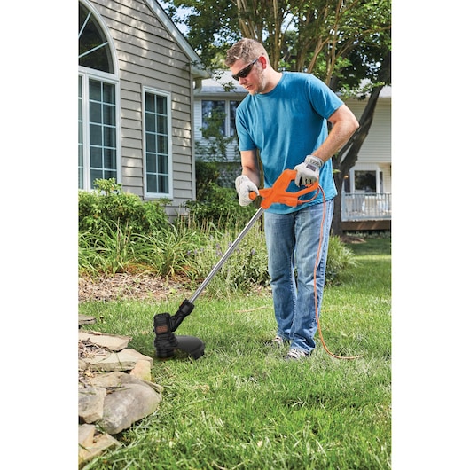 Photo of a person using a BLACK+DECKER BEST935 orange and black corded string trimmer on green lawn near a house.