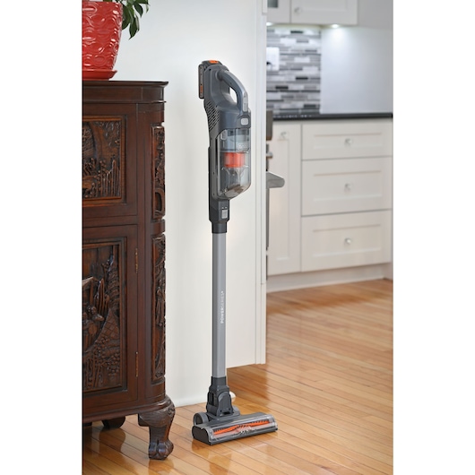 BLACKDECKER BHFEA18D1 cordless stick vacuum cleaner in gray and orange standing on hardwood floor near cabinet.