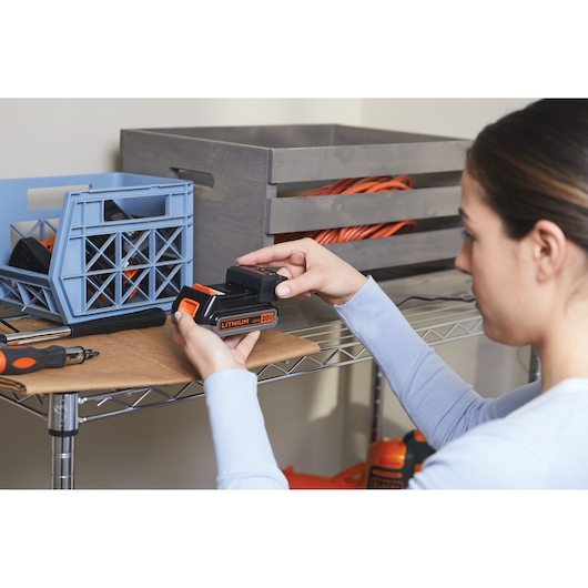 Photo of person holding BLACKDECKER 20V lithium battery BSV20PET in workshop on metal shelf with tools and storage boxes.