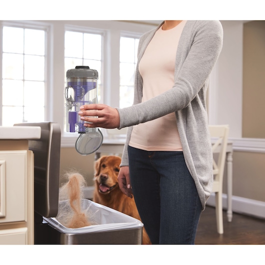 BLACKDECKER BSV20PET vacuum dirt canister being emptied into trash bin, dog in background, indoor setting.