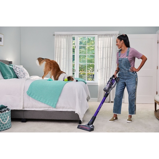 BLACKDECKER BSV20PET cordless stick vacuum held by a person cleaning bedroom carpet near dog and bed.