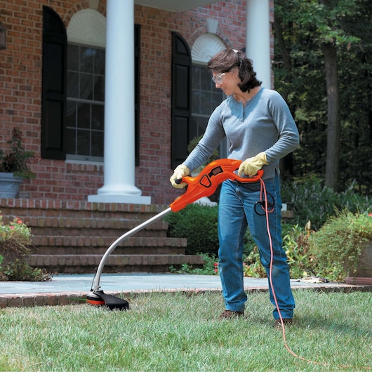 Photo of BLACKDECKER GH1100 string trimmer in use trimming grass near a brick house, orange body and curved shaft visible.