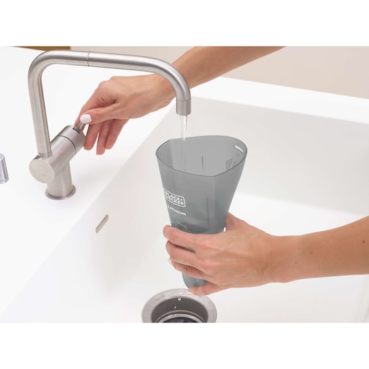 Photo of hands washing BLACK+DECKER HNV220V12 vacuum dustbin under a sink faucet in a white kitchen.