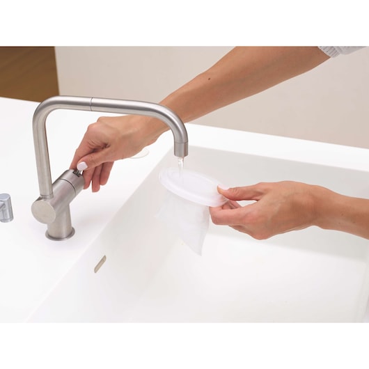 Photo of hands rinsing a BLACKDECKER HNV220V12 vacuum filter under a kitchen faucet in a white sink.