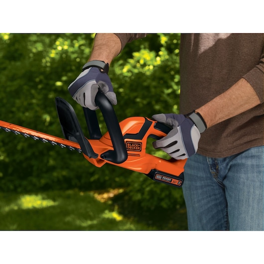 BLACKDECKER LHT2220 cordless hedge trimmer in use outdoors, orange body, operator wearing gloves.