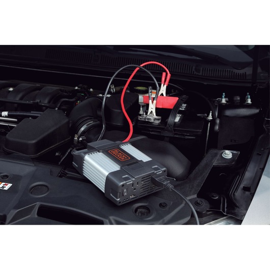 BLACKDECKER PI750LA power inverter connected to a car battery under hood, angled view, black and silver design.