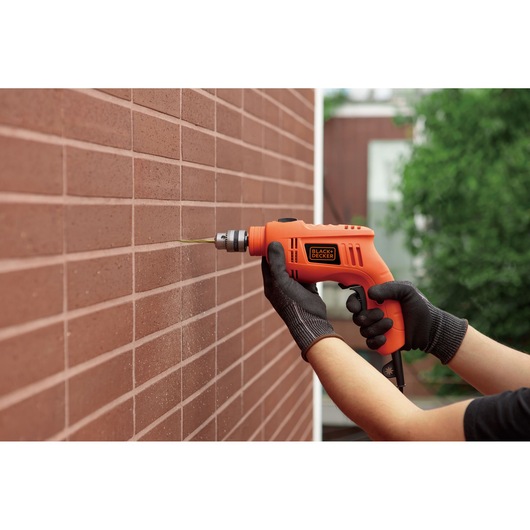 Photo of BLACKDECKER TB555 orange electric drill in use on a brick wall, held by gloved hands, outdoors.