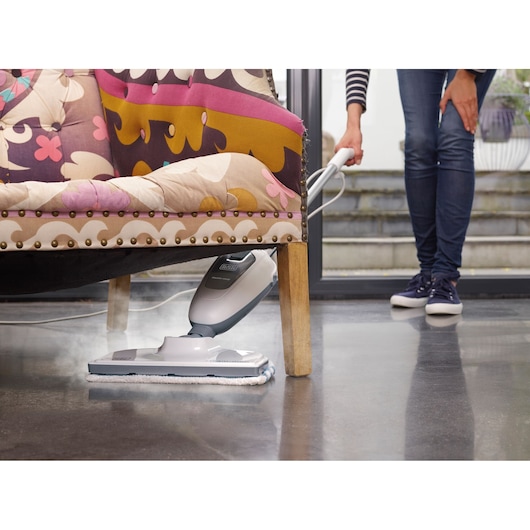 BLACKDECKER VL1300 steam mop cleaning under patterned sofa on a polished floor, handheld by a person.